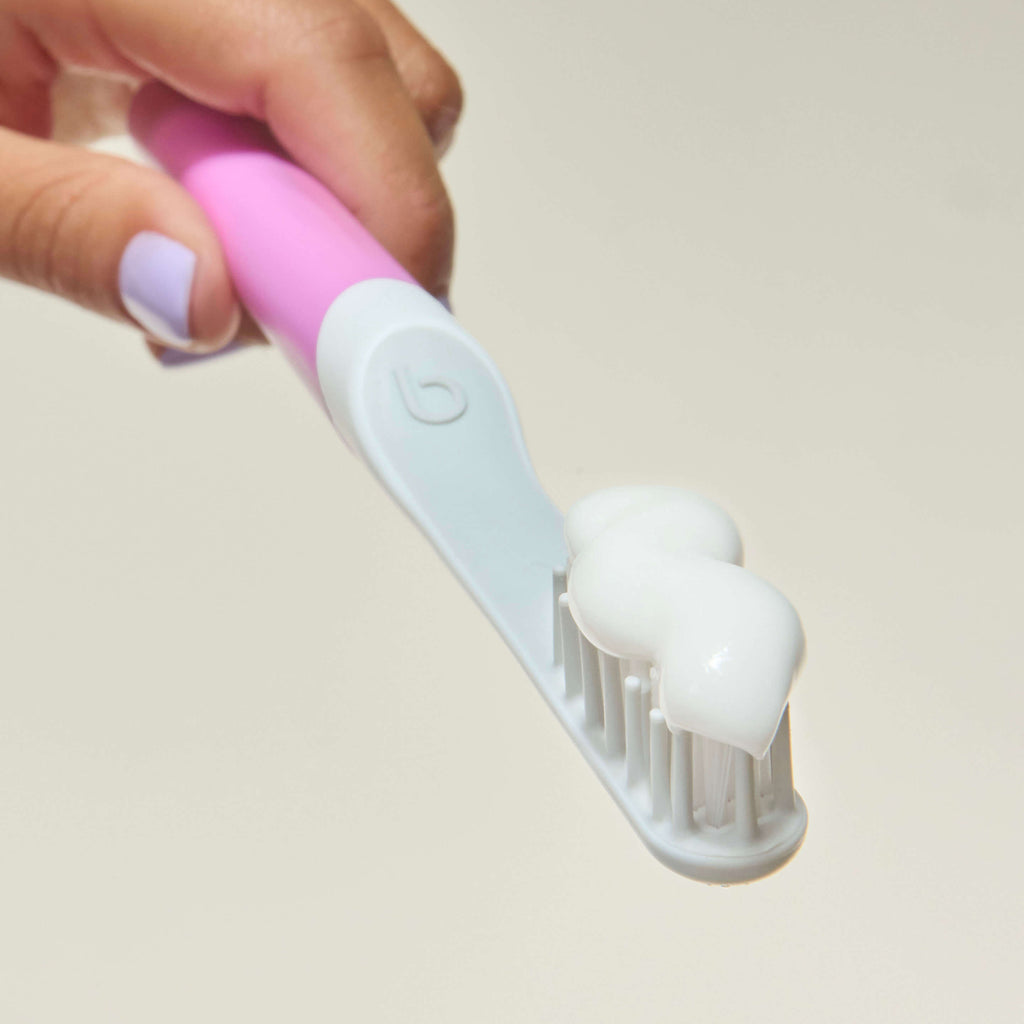 Close-up of a pink and light blue toothbrush head with soft white bristles, topped with a swirl of white toothpaste, held by a hand with lavender-painted nails.